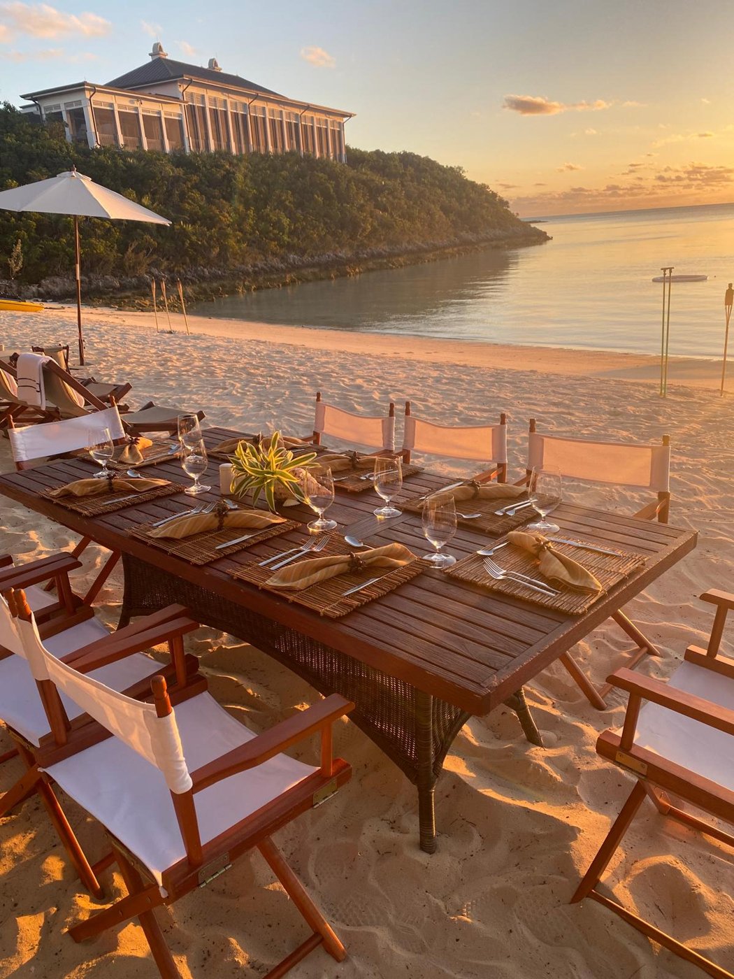 Beachfront dinner table set at sunset with glassware, flatware and folded serviettes